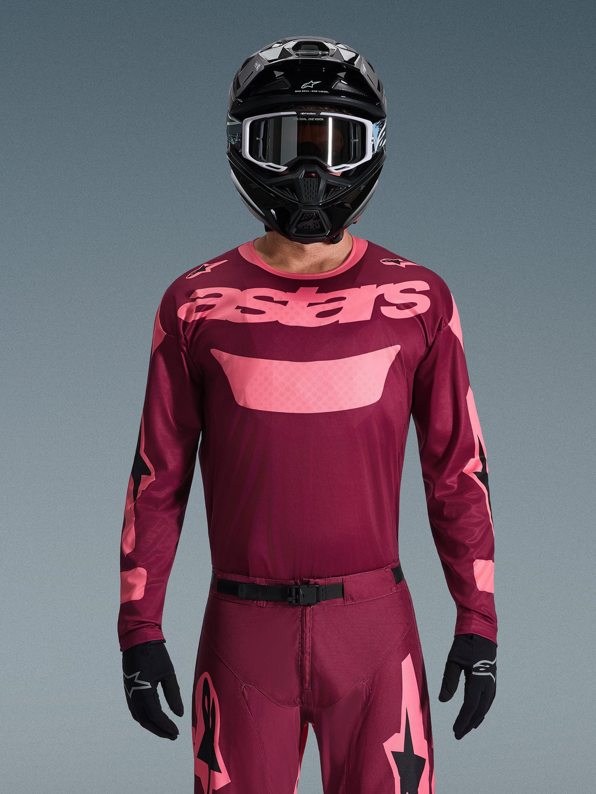 Motocrosskører iført Alpinestars 2026 Racer Riway Jersey i Dark Red Coral, med en vinrød krop og koralrosa 'astars'-branding på brystet, kombineret med matchende motocrossbukser, sort hjelm og sorte handsker, professionelt udstyrssæt til terrænkørsel.