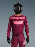 Motocrosskører iført Alpinestars 2026 Racer Riway Jersey i Dark Red Coral, med en vinrød krop og koralrosa 'astars'-branding på brystet, kombineret med matchende motocrossbukser, sort hjelm og sorte handsker, professionelt udstyrssæt til terrænkørsel.