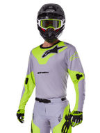 Alpinestars 2025 Racer Veil Gear Set, motocrosskører iført gråt og neongrønt udstyrssæt, lysegrå trøje og bukser med markante neongule paneler og sorte logoer, koordineret med matchende handsker og sort hjelm med neongrønne goggles, professionelt udstyr til terrænkørsel.