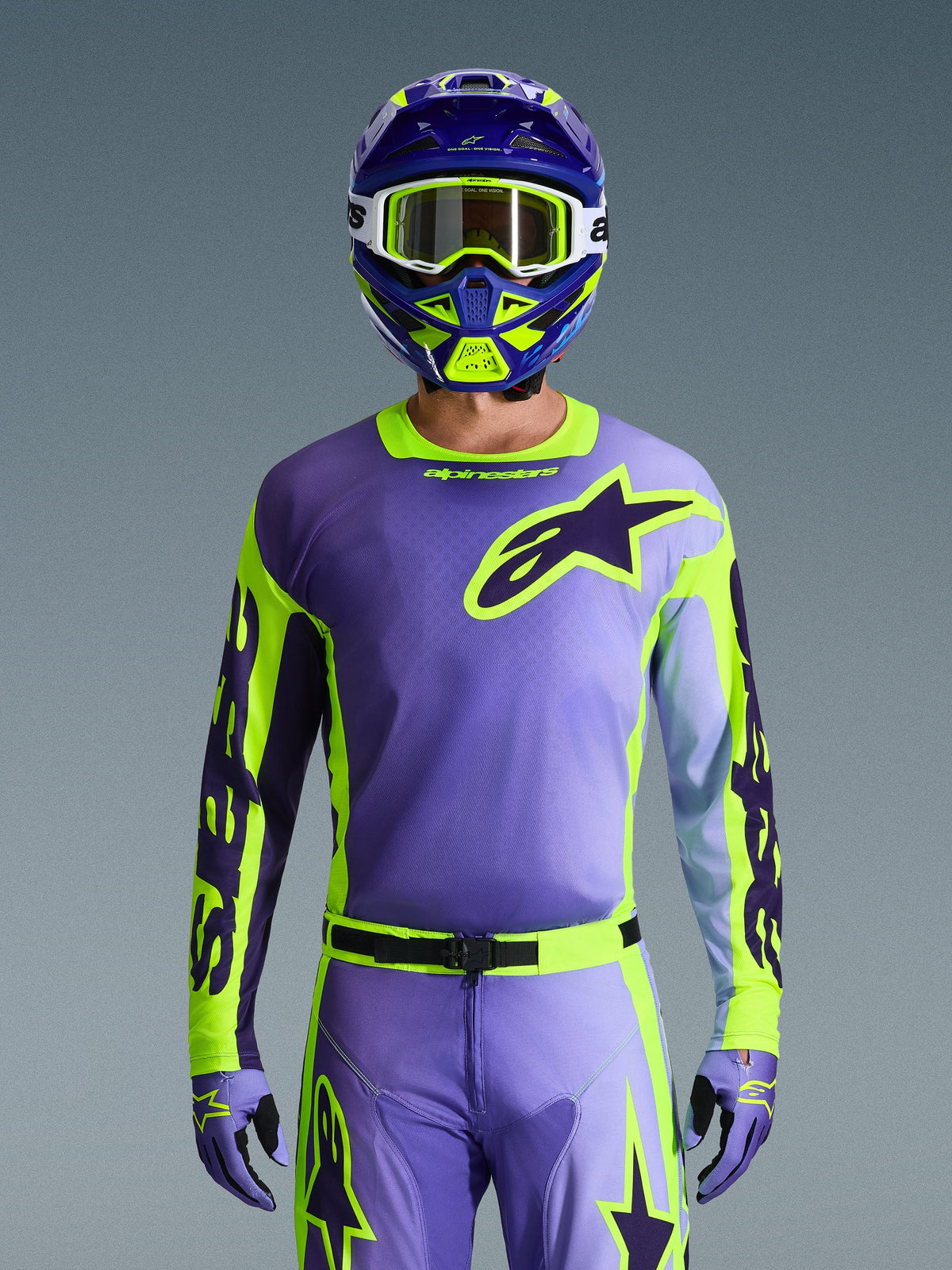 Alpinestars 2026 Racer Portl Jersey, motocross-trøje, lilla og neongul, båret med matchende motocross-bukser, handsker og hjelm, med et stort Alpinestars-stjernelogo på brystet, studiefoto mod en grå baggrund.