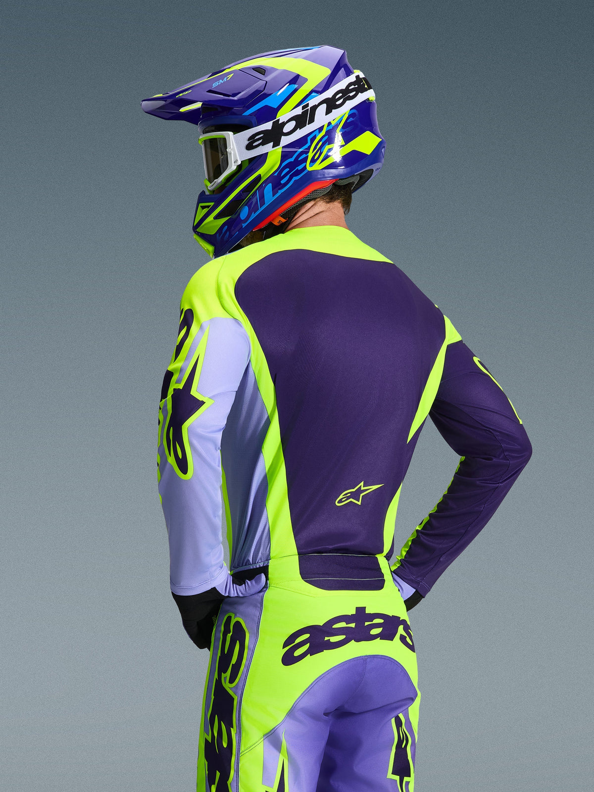 Motocrosskører set bagfra iført Alpinestars 2026 Racer Portl-trøje og matchende bukser, lilla og Yellow Fluo-farvekombination med lavendel-accenter, koordineret blå og gul hjelm, race-fit udstyr designet til off-road kørsel.