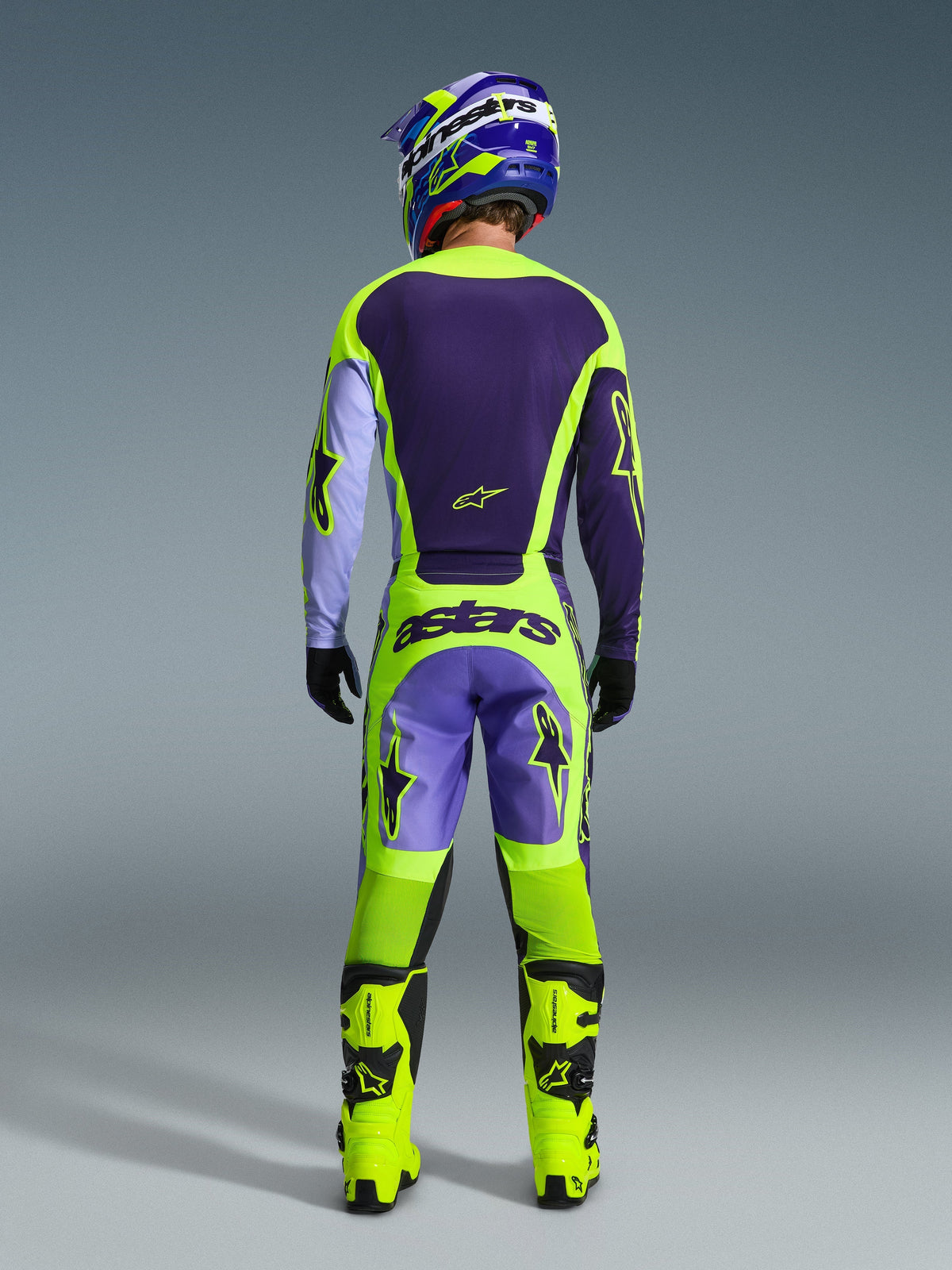 Motocross-kører set bagfra iført Alpinestars 2026 Racer Portl-trøje i lilla og neon-gul, matchende motocross-bukser, neon-gule støvler og blå og gul hjelm, komplet off-road udstyrssæt.