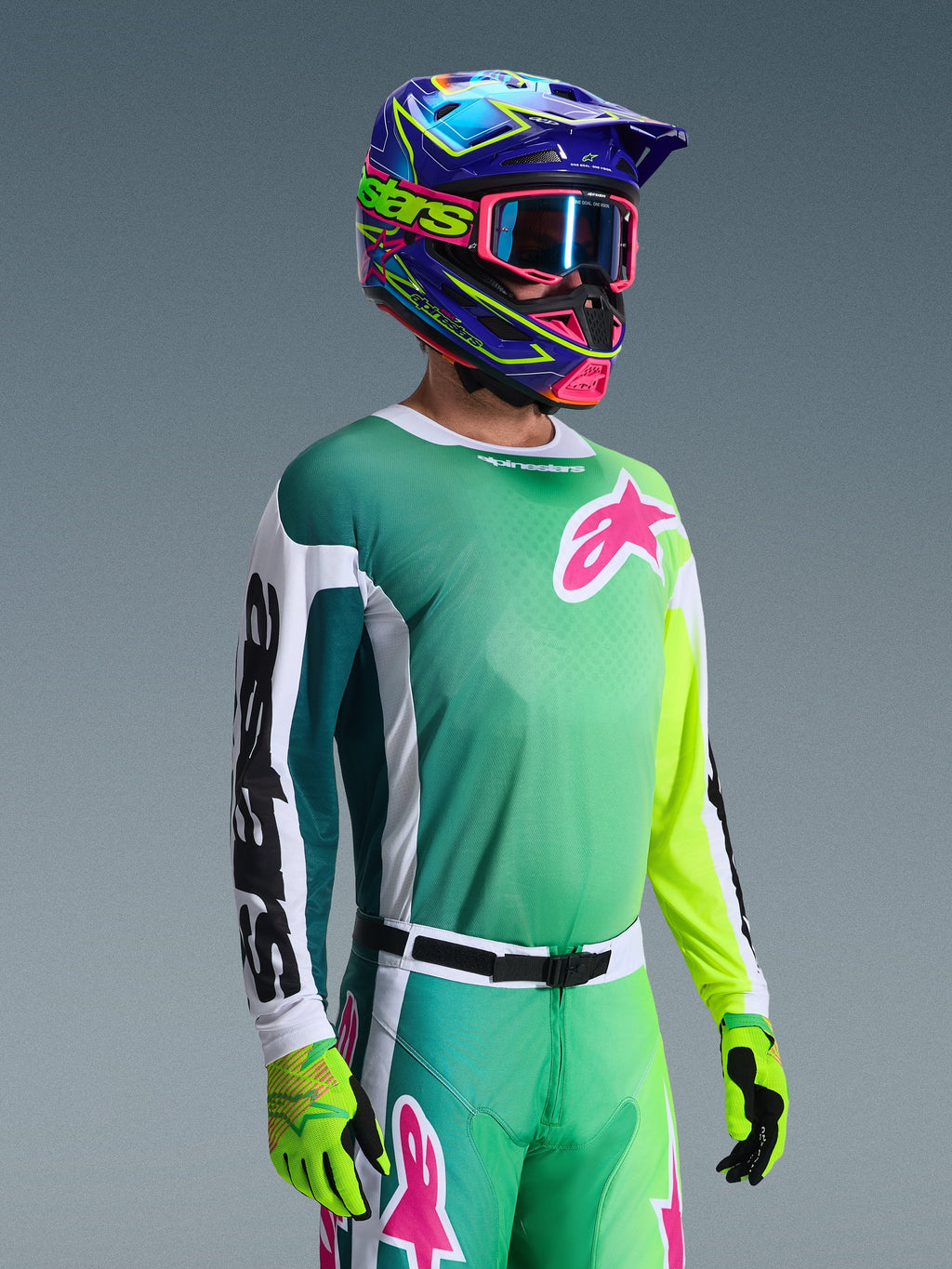 Alpinestars 2026 Racer Portl-trøje, motocross-trøje, grøn, hvid og sort, båret af en kører med matchende grønne bukser, fluorescerende handsker og en livlig blå og lilla hjelm med lyserøde goggles, med et lyserødt Astars-logo på brystet.