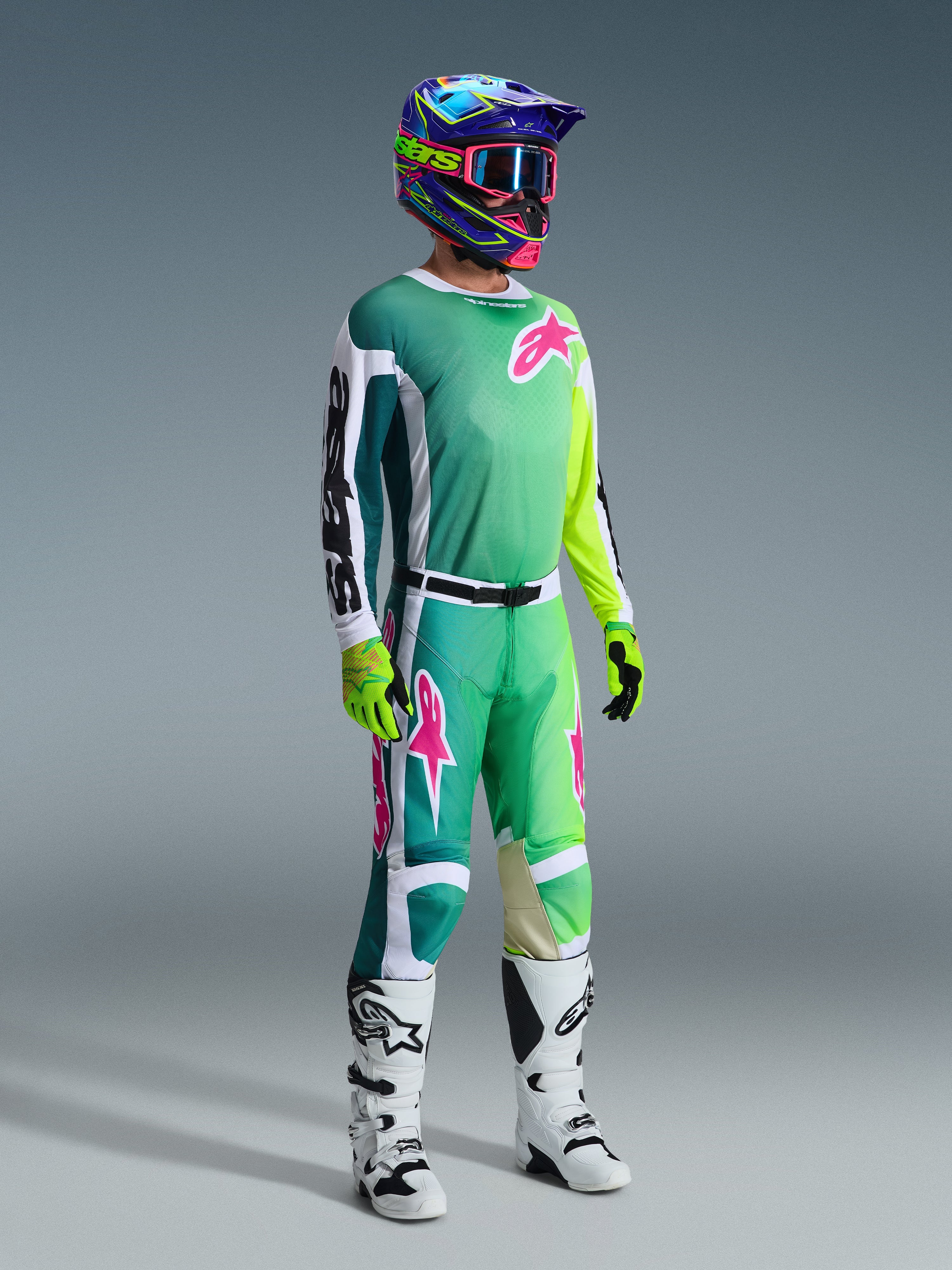 Alpinestars 2026 Racer Portl Gear Set, motocrosskører i jersey med gradient fra blågrøn til limegrøn med lyserøde logoer og hvide ærmer, matchende grønne og hvide bukser, farverig grafisk hjelm med lyserøde crossbriller, hvide støvler og fluorescerende gule handsker, professionelt off-road racerudstyr