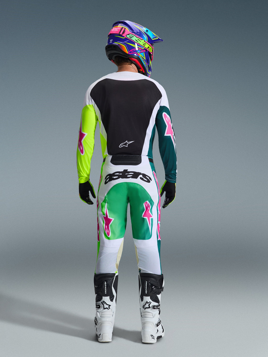 Set bagfra af motocross-kører iført Alpinestars 2026 Racer Portl-udstyrssæt, hvid trøje med sort rygpanel og neongule og mørkegrønne ærmer med lyserøde stjerner, matchende hvide bukser med grønne og blågrønne detaljer og lyserøde stjernelogoer, koordineret med en farverig hjelm, sorte handsker og hvide og sorte støvler.