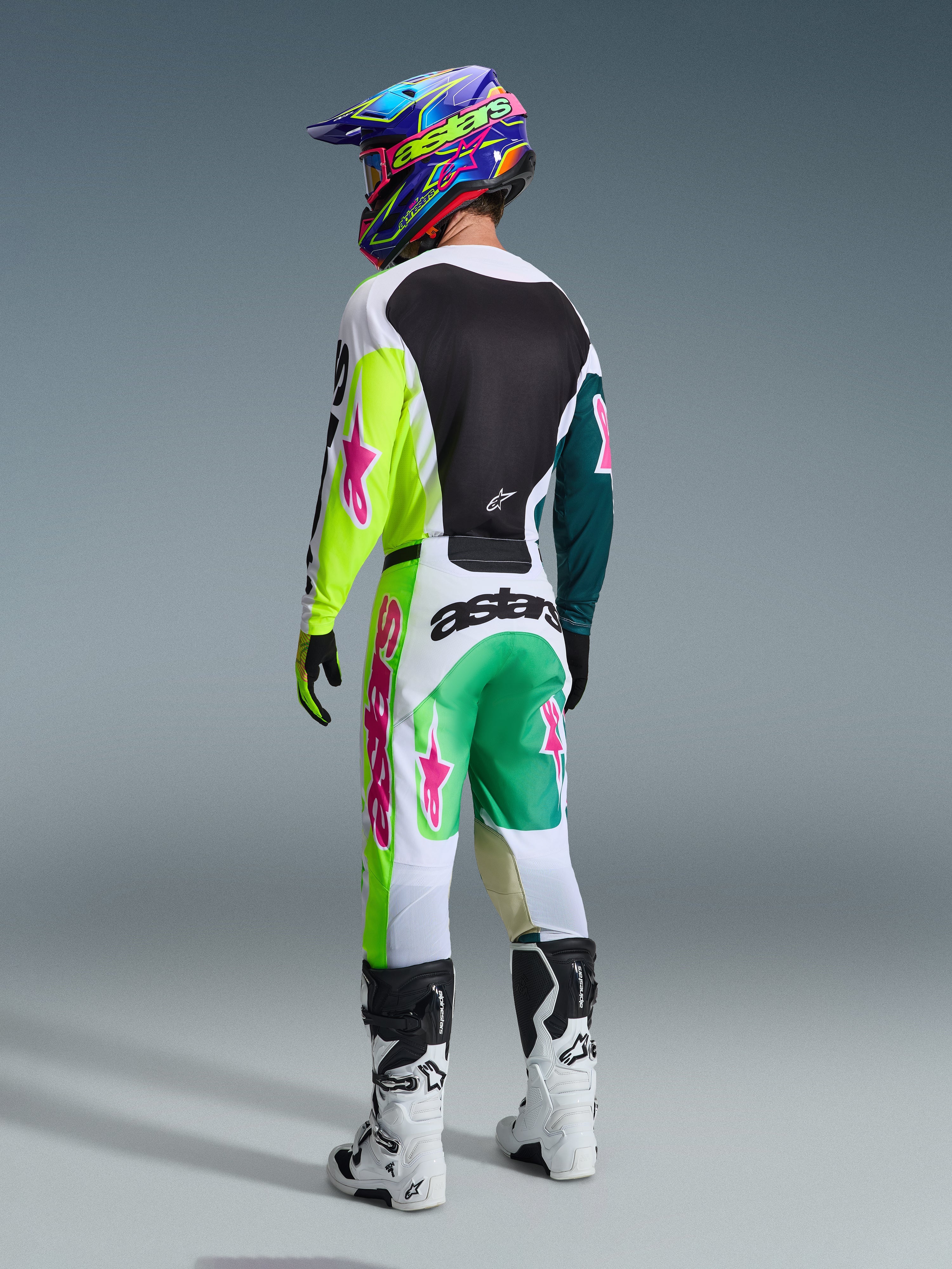 Motocrosskører set bagfra iført Alpinestars 2026 Racer Portl-udstyrssæt, trøje med neongule og blågrønne ærmer med lyserøde logoer, hvide og grønne bukser med markante lyserøde detaljer, farverig hjelm og hvide støvler, professionelt off-road racerudstyr