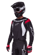 Motocross-kører iført Alpinestars 2025 Fluid Haul udstyrssæt, sort jersey med røde skulderpaneler og hvid grafik, matchende sorte bukser med hvide stjernelogoer, rød-sorte handsker, sort hjelm med hvide detaljer og goggles, koordineret motocross-beklædning.