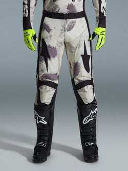 Alpinestars 2026 Racer Lahnd Pants, Motocross Pants, Sand Camo pattern with gray and purple abstract graphics, worn with neon yellow gloves and black boots, featuring black star logos on the thighs and a black waist belt, durable off-road racing gear