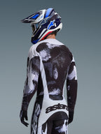 Alpinestars 2026 Racer Lahnd Jersey i Iron Camo, set bagfra af en motocrosskører i et sort, hvidt og gråt udstyrssæt med abstrakt mønster, inklusiv matchende bukser, sorte handsker og en blå racerhjelm med crossbriller.