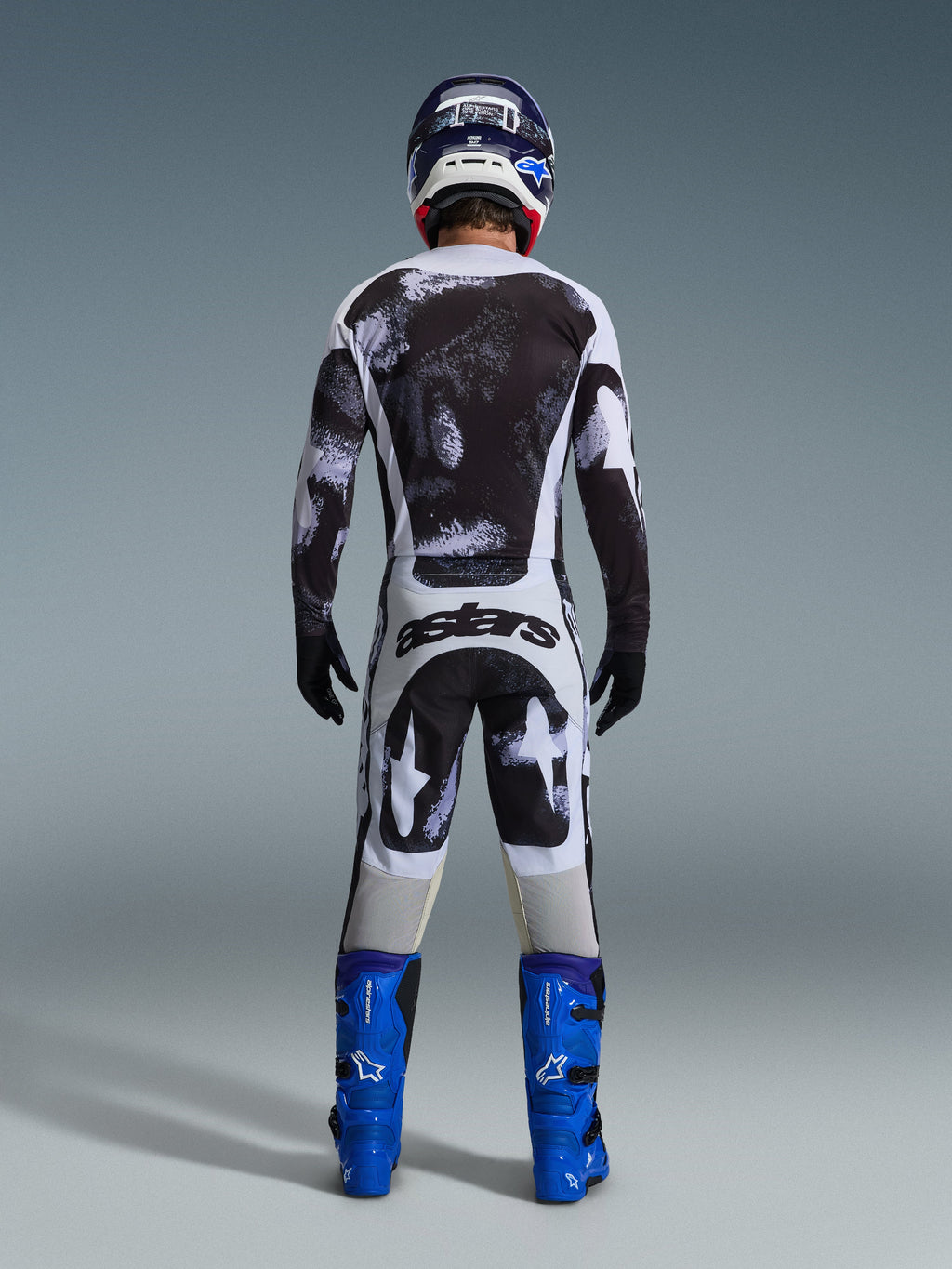 Motocrosskører set bagfra iført Alpinestars 2026 Racer Lahnd-udstyrssæt, sort og hvid trøje og bukser med abstrakt mønster, 'astars'-branding på bagen, kombineret med blå støvler og en matchende hjelm, professionel beklædning til terrænkørsel.