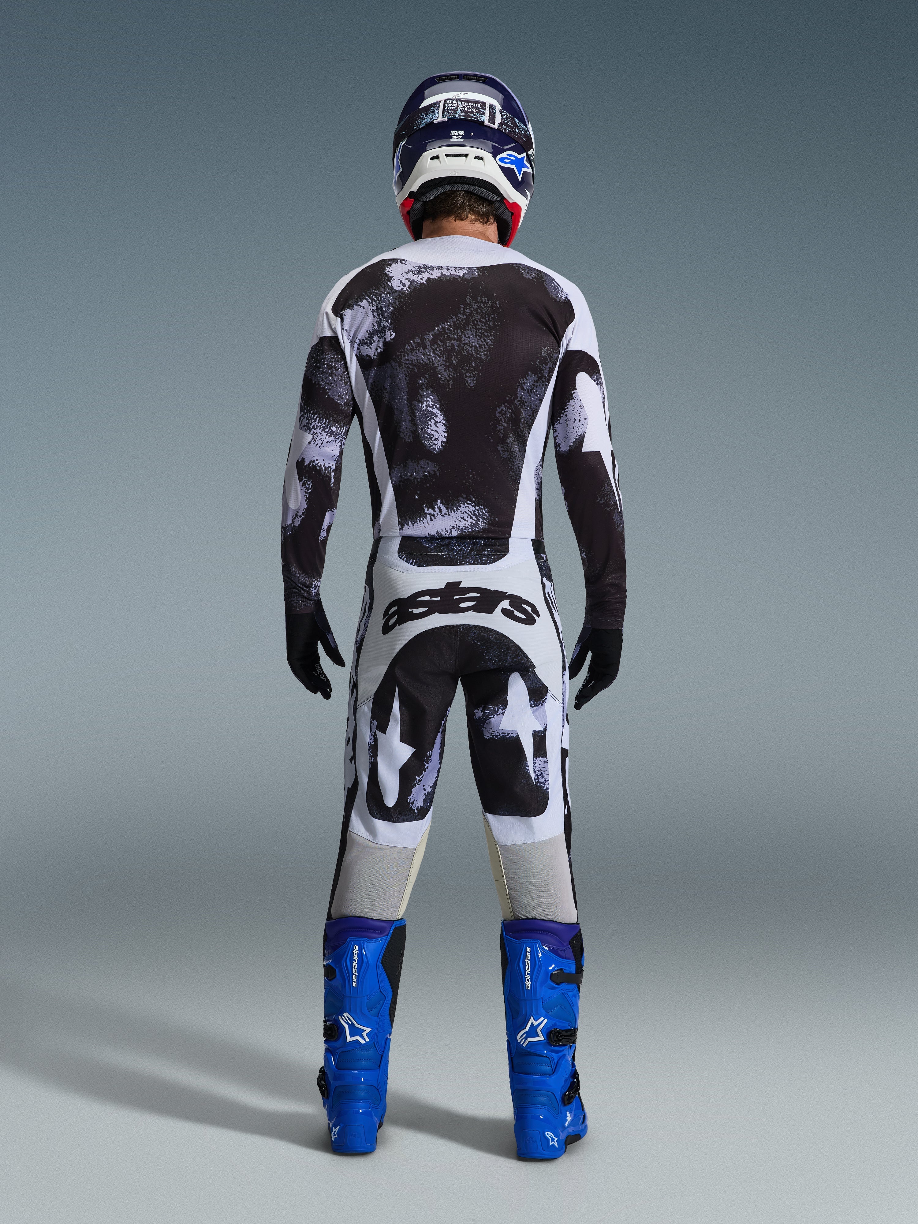 Motocrosskører set bagfra iført Alpinestars 2026 Racer Lahnd-udstyrssæt, sort og hvid trøje og bukser med abstrakt mønster, 'astars'-branding på bagen, kombineret med blå støvler og en matchende hjelm, professionel beklædning til terrænkørsel.