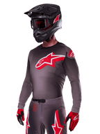 Alpinestars 2025 Supertech Lipan Gear Set, motocross-kører iført mørkegrå trøje og bukser med røde og hvide detaljer, stort rødt Alpinestars-logo på brystet, laserskårne perforeringer til ventilation, matchende sort hjelm, røde goggles og rød-sorte handsker.