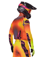 Motocross-kører set bagfra fra siden iført Alpinestars 2025 Supertech Lipan-udstyrssæt, med en gul, orange og rød gradient med sort og lyserød branding, sort hjelm med lyserøde detaljer og orange crossbriller, professionel beklædning til terrænkørsel.