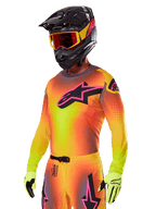 Motocrosskører iført Alpinestars 2025 Supertech Lipan-udstyrssæt, med en jersey med gul og orange farveovergang og et lyserødt og sort Astars-logo, neongule handsker og en sort hjelm med røde briller, professionelt terrænkørselsudstyr.