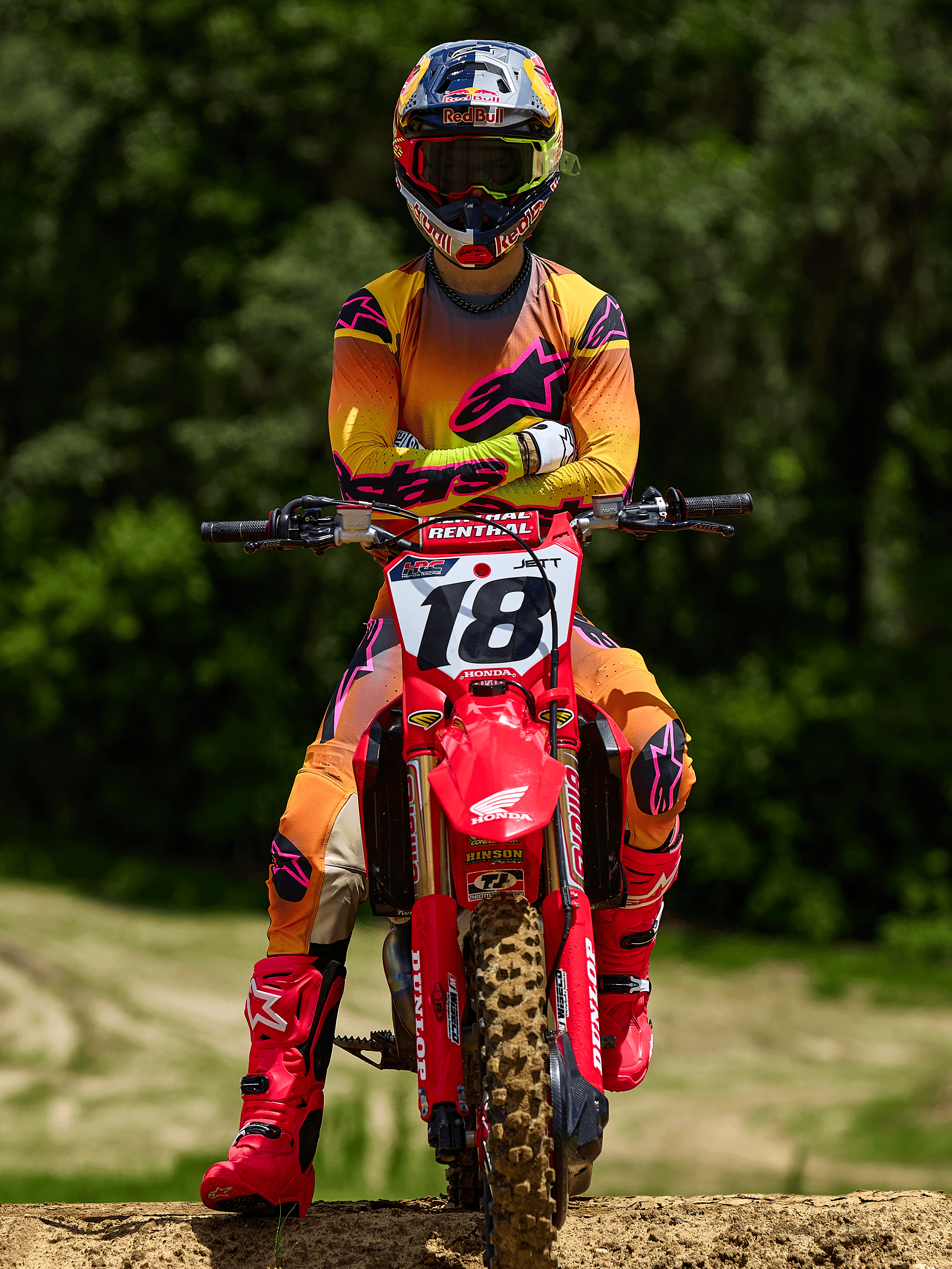 Motocross rider sitting on a red Honda dirt bike with racing number 18, wearing Alpinestars 2025 Supertech Lipan Jersey and matching pants in Yellow and Pink, bright pink Alpinestars boots, Red Bull helmet, professional racing gear for off-road performance.