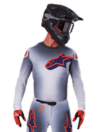 Alpinestars 2025 Supertech Lipan Jersey, motocross racertrøje, grå med orange og mørkeblå detaljer, langærmet performance-top med perforerede ventilationsdetaljer, vist med sort hjelm og røde handsker, strømlinet racerdesign