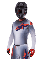 Motocrosskører iført Alpinestars 2025 Supertech Lipan-udstyrssæt, grå jersey og bukser med navy og orange detaljer, mat sort hjelm med goggles og rød-sorte handsker, atletisk præstationsudstyr til terrænkørsel