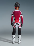 Alpinestars 2026 Supertech AFD Vista Gear Set, set bagfra af en motocrosskører i professionelt racerudstyr, vinrød og rød jersey med hvide og sorte detaljer, matchende bukser med markant grafik, hvide støvler og sort hjelm, koordineret motocross-raceroutfit.