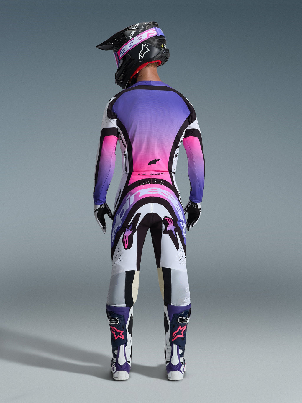 Motocrosskører set bagfra iført 2026 Supertech AFD Vista Jersey, hvid, lilla og sort med pink gradient-detaljer, matchende motocrossbukser, sort hjelm med lyserøde goggles og hvide og lilla støvler, komplet professionelt racersæt