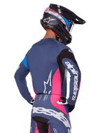 Motocrosskører set bagfra iført Alpinestars 2025 Techstar Dreem-udstyrssættet i grå, bestående af en blågrå jersey og bukser med lyserøde og sorte paneler, hvide Alpinestars-logoer på ærmerne og ryggen samt en matchende sort hjelm med blå og lyserød grafik.