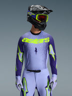Alpinestars 2026 Supertech Scenz Gear Set, motocross-udstyr, frontal visning af kører, lyslilla og mørkeblå trøje med neongul 'astars'-branding, matchende lyslilla bukser og handsker med neon-detaljer, sort hjelm med neongule briller, studiofoto mod grå baggrund