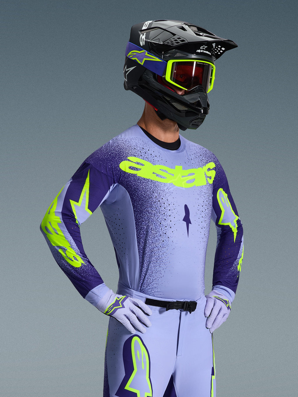 Motocross-kører iført 2026 Alpinestars Supertech Scenz-trøje, lilla og gul fluo, matchende lilla bukser med fluo-detaljer, sort hjelm med gule briller og lilla racinghandsker, koordineret professionelt udstyr til terrænkørsel