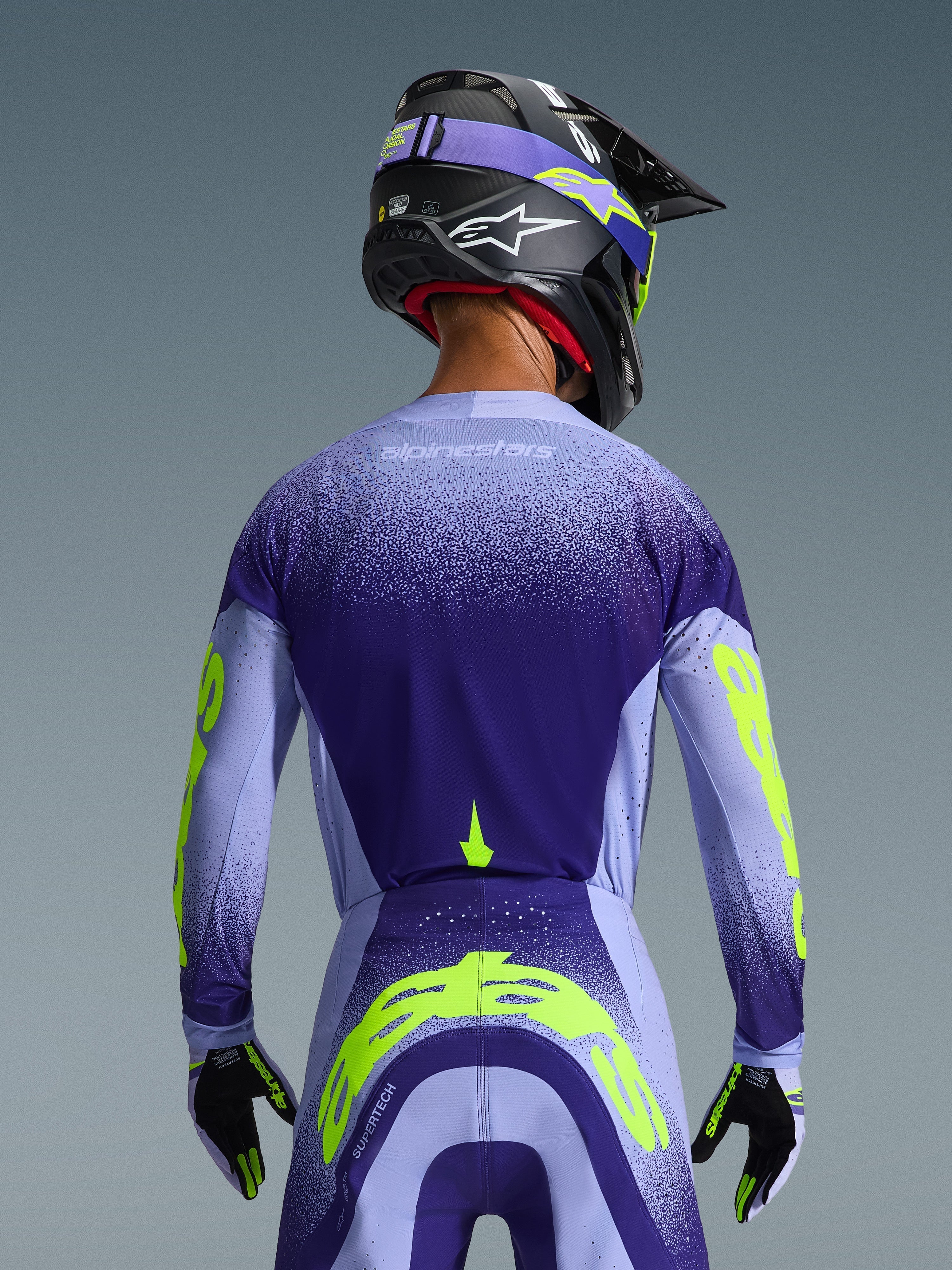 Alpinestars 2026 Supertech Scenz trøje i lilla og lyseblå med fluoriserende gule detaljer, set bagfra på kører iført matchende motocrossbukser, sort hjelm og handsker, højtydende atletisk udstyr til terrænkørsel