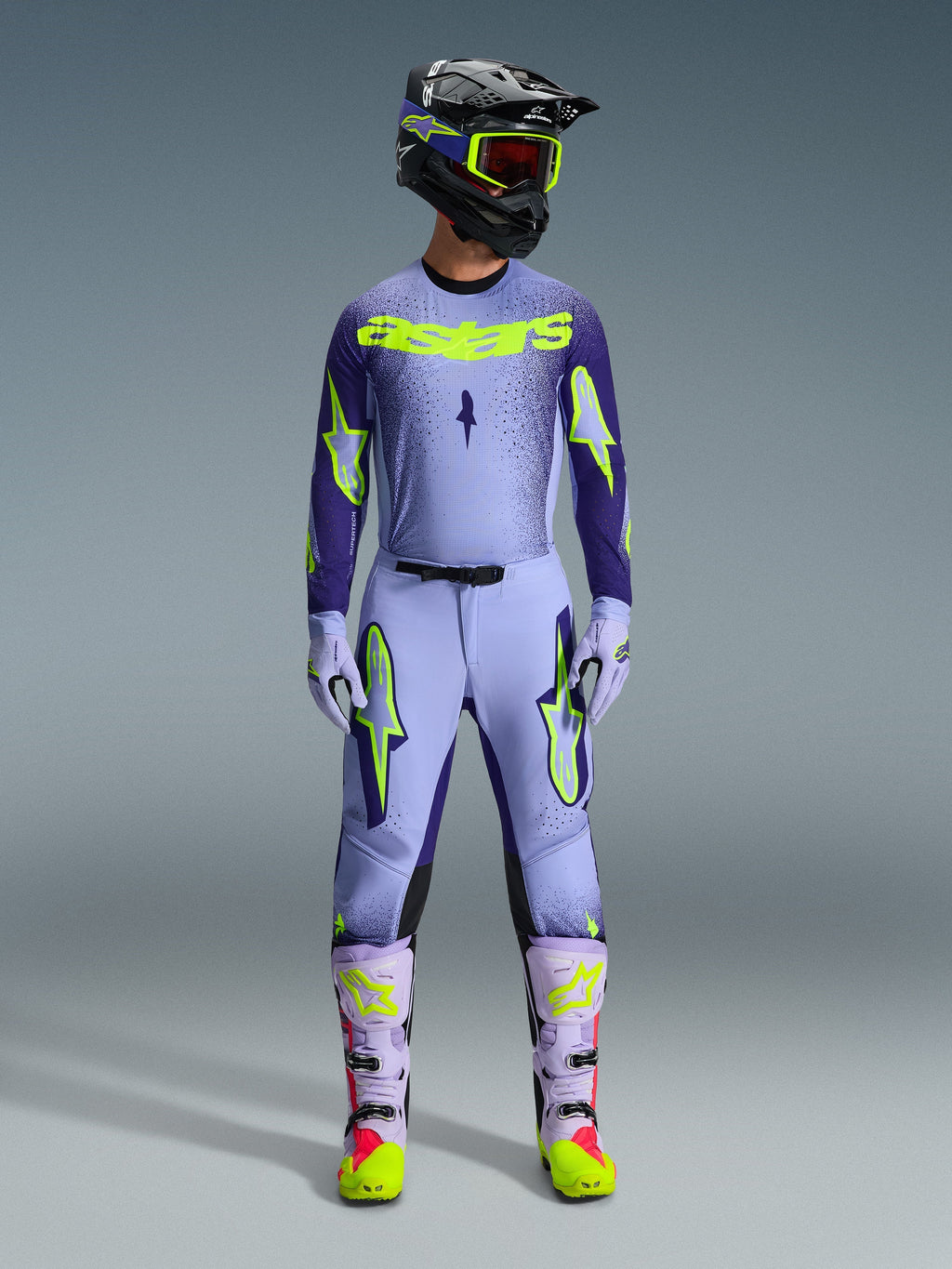 Motocross-kører iført komplet Alpinestars 2026 Supertech Scenz-racerudstyr, lilla og gul fluo-trøje med matchende bukser, sort hjelm med gule goggles, lyslilla og neon-støvler, professionelt outfit med atletisk pasform til terrænkørsel