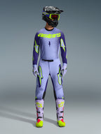 Motocross-kører iført komplet Alpinestars 2026 Supertech Scenz-racerudstyr, lilla og gul fluo-trøje med matchende bukser, sort hjelm med gule goggles, lyslilla og neon-støvler, professionelt outfit med atletisk pasform til terrænkørsel