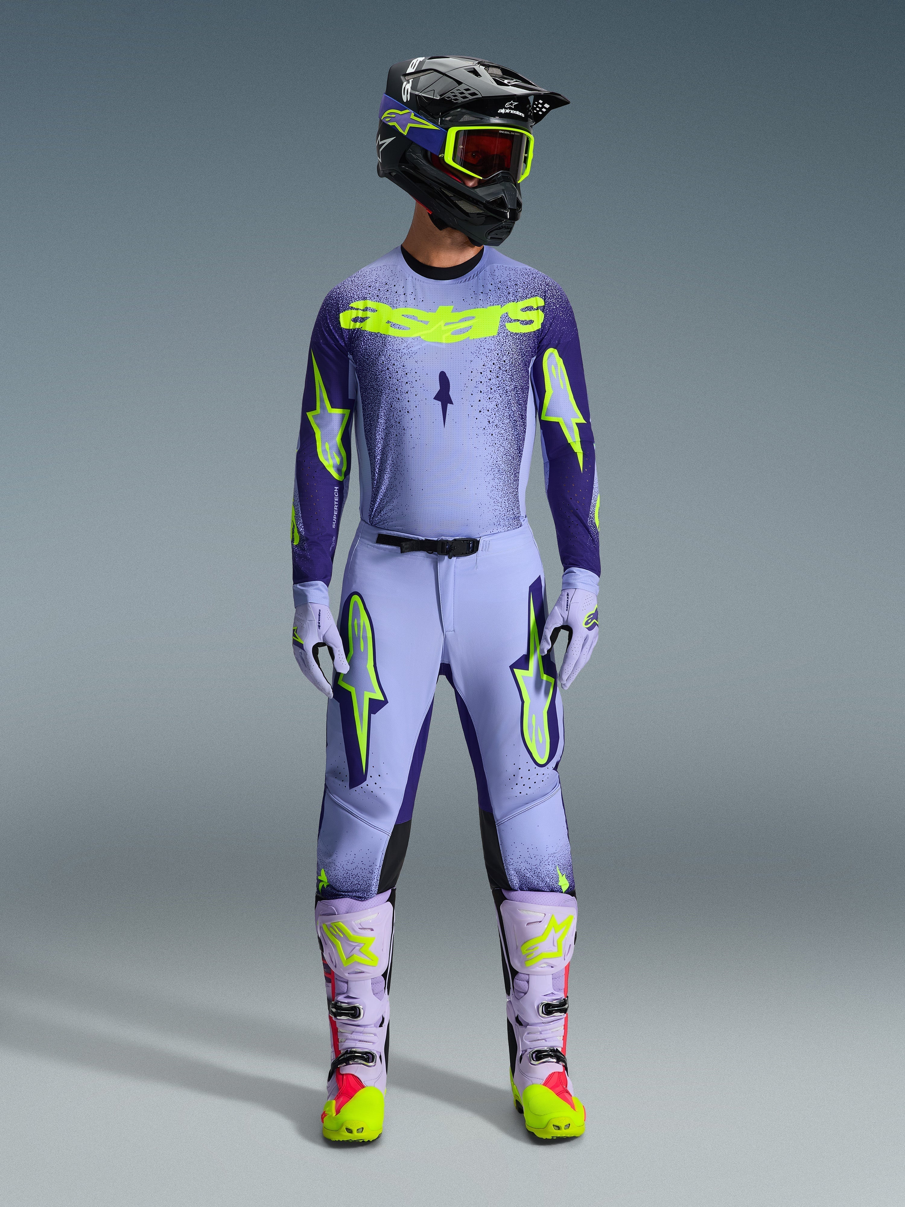 Motocross-kører iført komplet Alpinestars 2026 Supertech Scenz-racerudstyr, lilla og gul fluo-trøje med matchende bukser, sort hjelm med gule goggles, lyslilla og neon-støvler, professionelt outfit med atletisk pasform til terrænkørsel