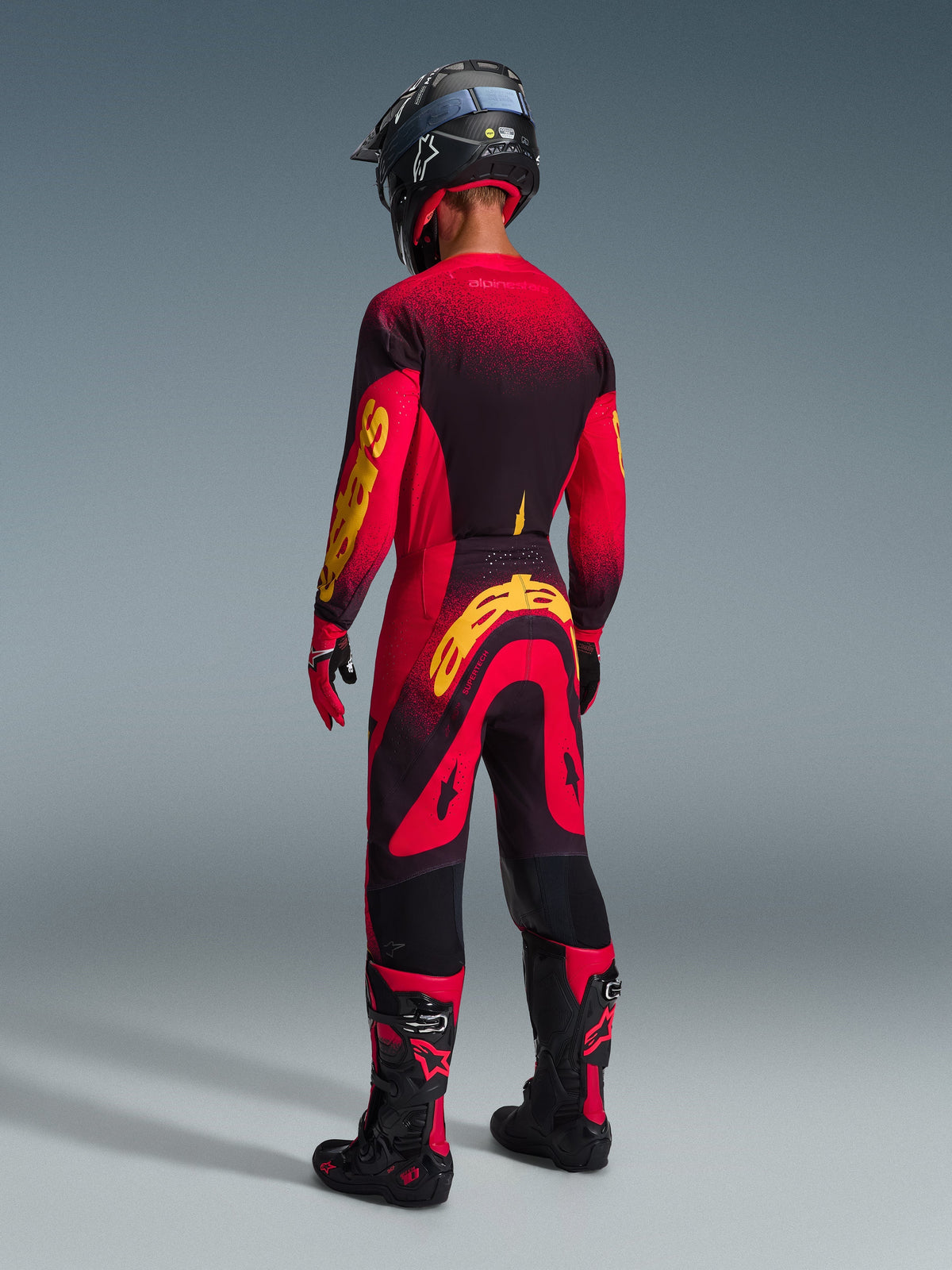 Motocrosskører set bagfra iført komplet racerudstyr, rød trøje med sorte og gule detaljer, matchende røde og sorte bukser med et stort gult Alpinestars-logo, sort hjelm, sort-røde støvler og rød-sorte handsker, koordineret sæt til professionel terrænkørsel