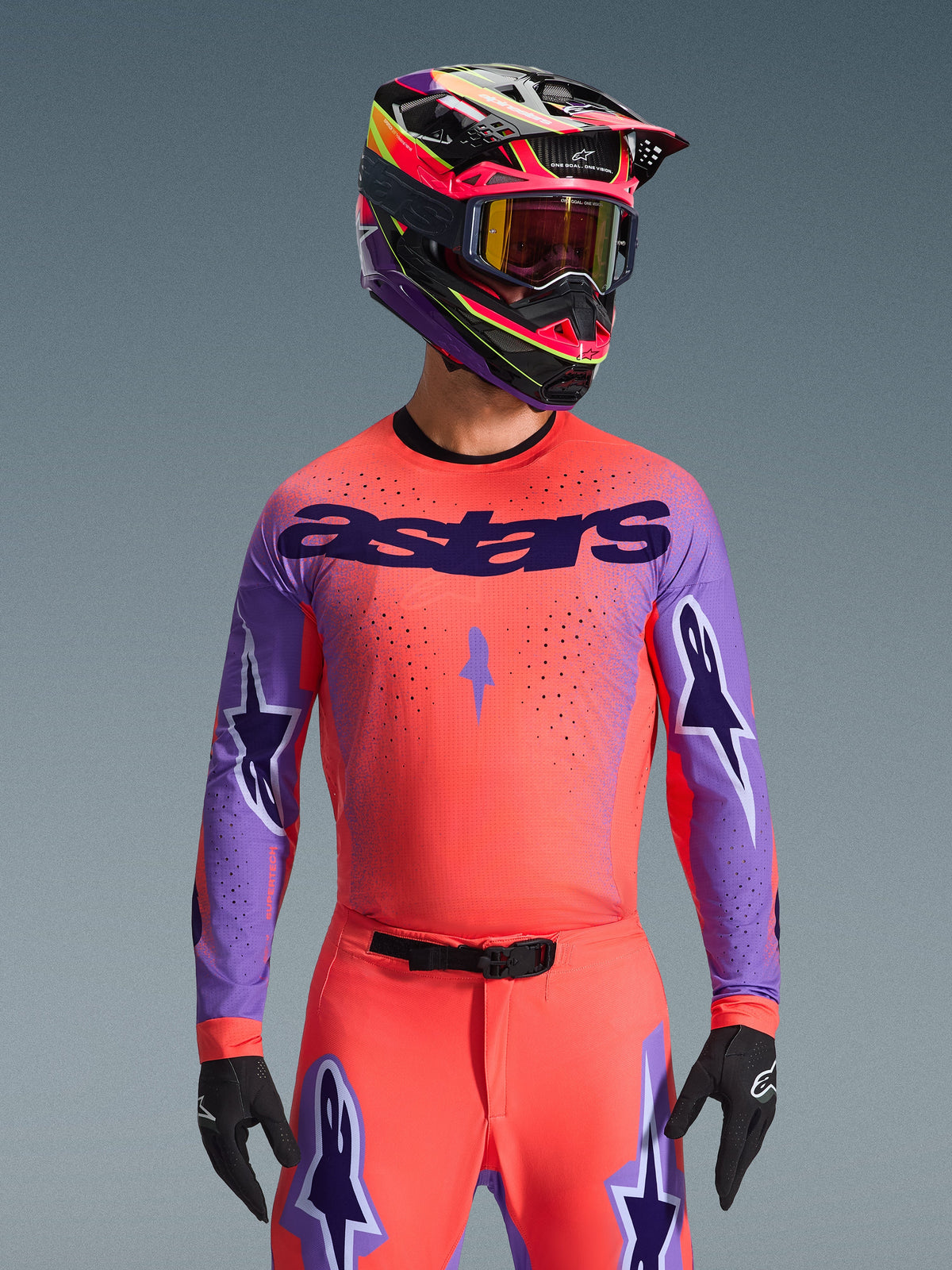 Alpinestars 2026 Supertech Scenz Jersey, motocross racingtrøje, Hot Coral og lilla, med stort 'astars'-logo og stjernedesign på ærmerne, båret med matchende bukser og en multifarvet hjelm, højtydende off-road gear