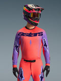 Alpinestars 2026 Supertech Scenz Jersey, motocross racingtrøje, Hot Coral og lilla, med stort 'astars'-logo og stjernedesign på ærmerne, båret med matchende bukser og en multifarvet hjelm, højtydende off-road gear