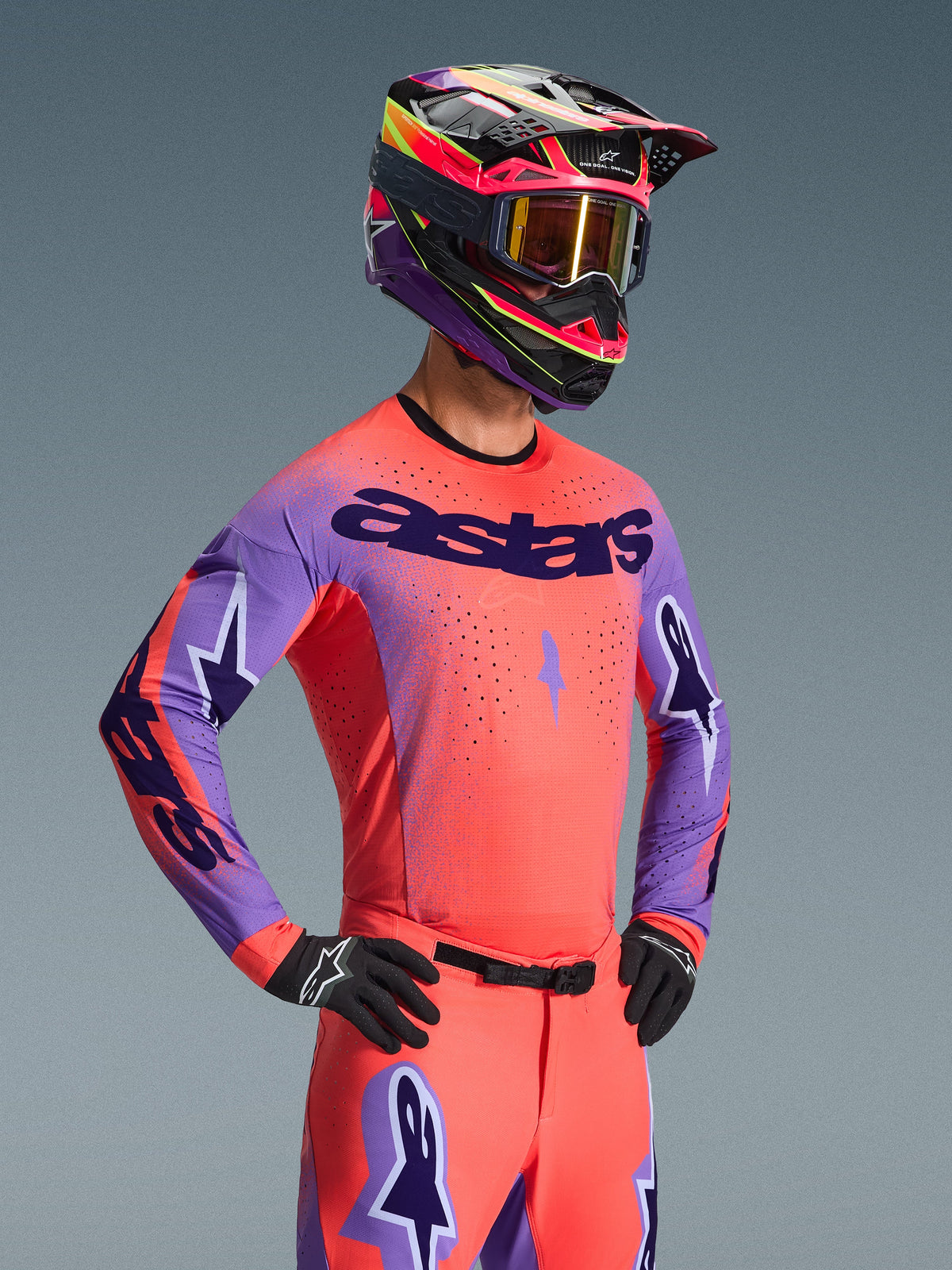 Motocross-kører iført Alpinestars 2026 Supertech Scenz-trøje, Hot Coral og lilla med markant 'astars'-branding på brystet, matchende udstyr inklusiv koralfarvede bukser og en koordineret lilla og koralfarvet hjelm, med laserskårne perforeringer til højtydende terrænkørsel.