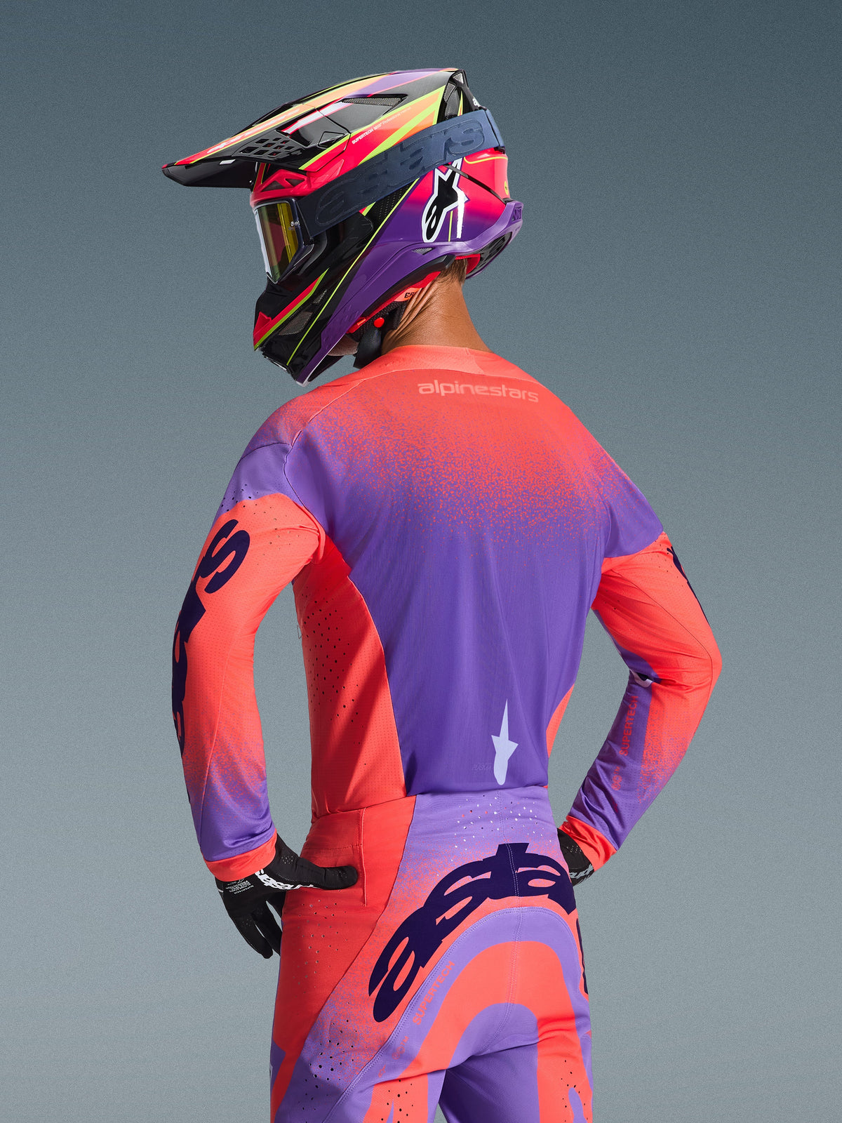 Motocrosskører set bagfra iført 2026 Alpinestars Supertech Scenz trøje i Hot Coral og lilla, med et livligt nistret gradientdesign, kombineret med matchende motocrossbukser og en farverig racerhjelm, professionelt udstyr til terrænkørsel.