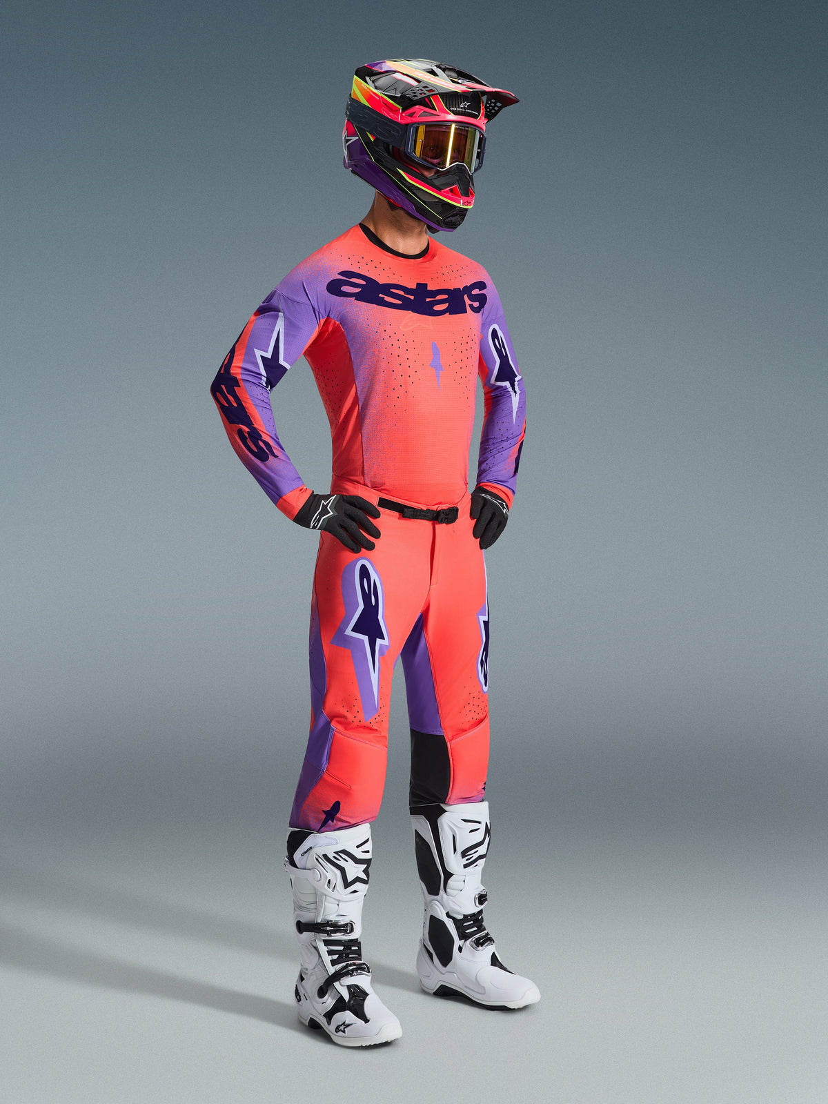Motocrosskører iført komplet Alpinestars 2026 Supertech Scenz-udstyrssæt, Hot Coral-jersey med lilla ærmer og 'astars'-branding, matchende coral-bukser med lilla detaljer, flerfarvet hjelm med guldtonede goggles, hvide og sorte støvler, sorte handsker, koordineret raceroutfit på professionelt niveau til terrænkørsel