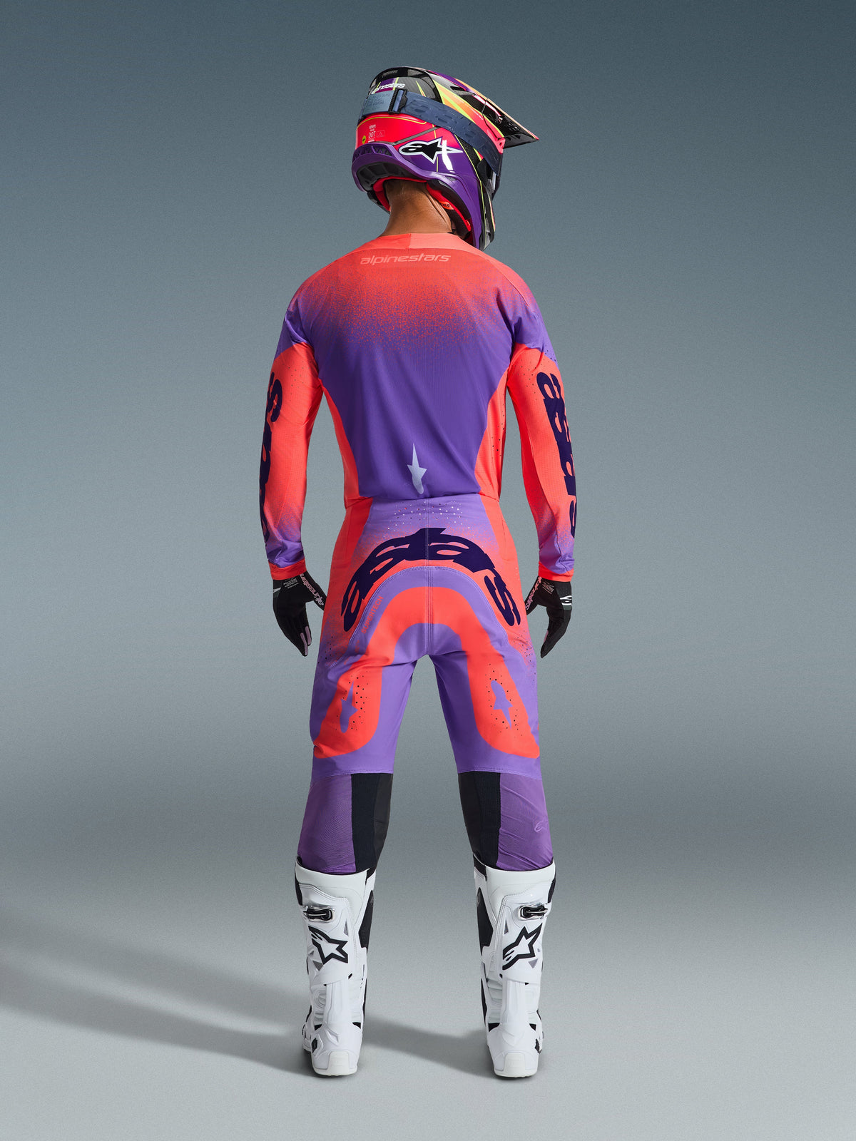 Motocrosskører set bagfra iført 2026 Supertech Scenz-udstyrssæt i Hot Coral Purple, strålende trøje og bukser med gradueret farveovergang, med Alpinestars-logoer, matchende hjelm, hvide og sorte støvler samt sorte handsker, professionelt udstyr til elite off-road racing.