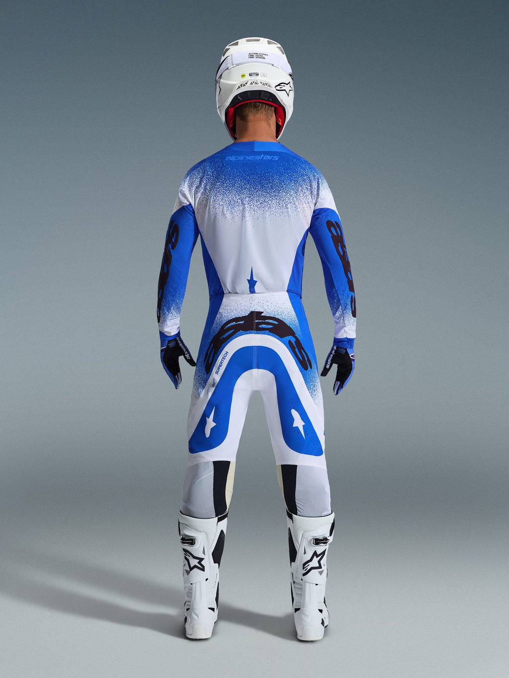 Motocrosskører set bagfra iført Alpinestars 2026 Supertech Scenz-udstyrssæt i Ucla Blue White, blå og hvid jersey med sort branding, matchende bukser, hvid hjelm og hvide støvler, koordineret professionelt racerudstyr til off-road motocross