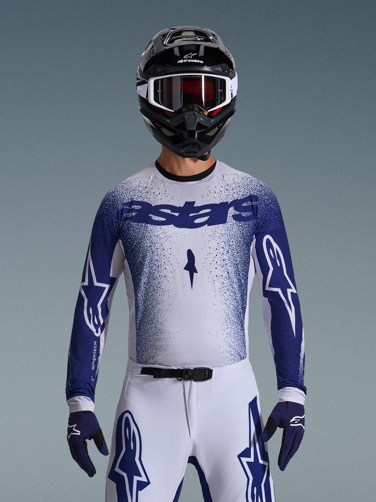 Motocross rider wearing the Alpinestars 2026 Supertech Scenz Jersey, Light Gray and Dark Blue with a speckled gradient design and bold 'astars' branding. Worn with matching white motocross pants, dark blue gloves, and a black helmet with goggles, coordinated outfit for professional off-road racing.