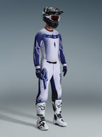 Motocross-kører iført komplet professionelt racerudstyr, 2026 Supertech Scenz-trøje i lysegrå og mørkeblå, matchende motocrossbukser med stjernegrafik, sort hjelm med røde goggles, hvide og sorte performance-støvler, koordinerede blå og hvide handsker til terrænkørsel
