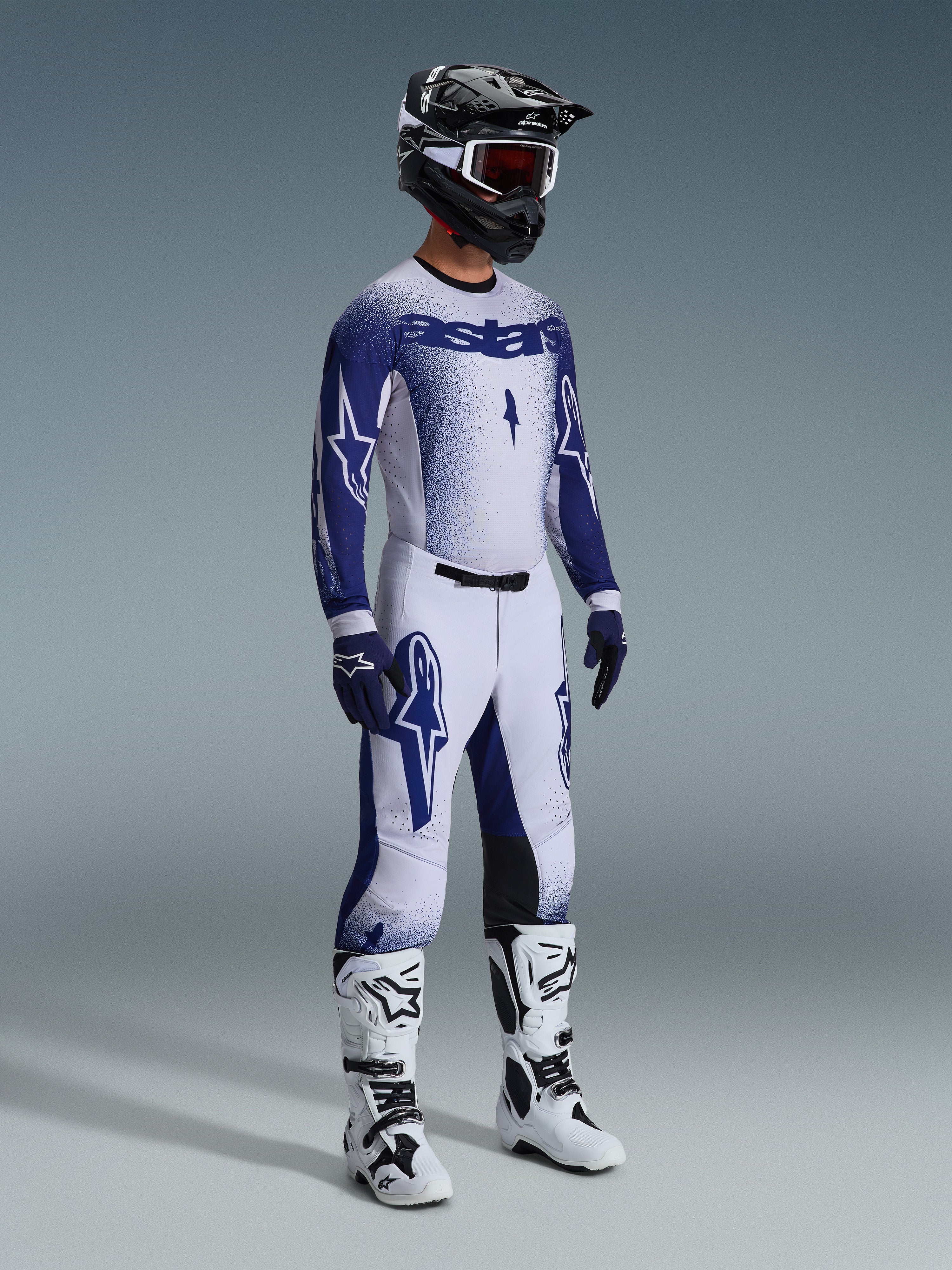 Motocross-kører iført komplet professionelt racerudstyr, 2026 Supertech Scenz-trøje i lysegrå og mørkeblå, matchende motocrossbukser med stjernegrafik, sort hjelm med røde goggles, hvide og sorte performance-støvler, koordinerede blå og hvide handsker til terrænkørsel