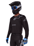 Motocross-kører iført Alpinestars 2025 Racer Graphite-udstyrssæt, sort jersey med grå Alpinestars-logoer, matchende sorte bukser med grå detaljer, sort hjelm med blå og hvide goggles, blå og grå handsker, strømlinet teknisk outfit til terrænkørsel