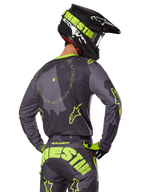 Alpinestars 2025 Racer Hollow Gear Set, motocross-kører set bagfra, grå trøje med fluorescerende gul cirkulær tekst og stjernelogoer, matchende grå og gule bukser med iøjnefaldende branding over sædet, sort og limegrøn hjelm, koordineret outfit til off-road racing