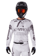 Alpinestars 2025 Racer Hollow Jersey, motocross-trøje, hvid og sort, båret med matchende hvide MX-bukser og en sort hjelm med orange goggles, med markant Alpinestars-branding og globusgrafik, professionelt off-road racerudstyr
