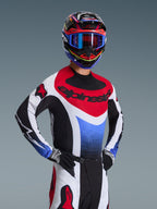 Motocrosskører iført Alpinestars 2026 Techstar Knif-trøje i rød, hvid og blå, med sort branding på brystet, kombineret med matchende motocrossbukser, en koordineret racerhjelm med blå goggles og sort-hvide handsker, professionelt off-road-udstyr til konkurrence