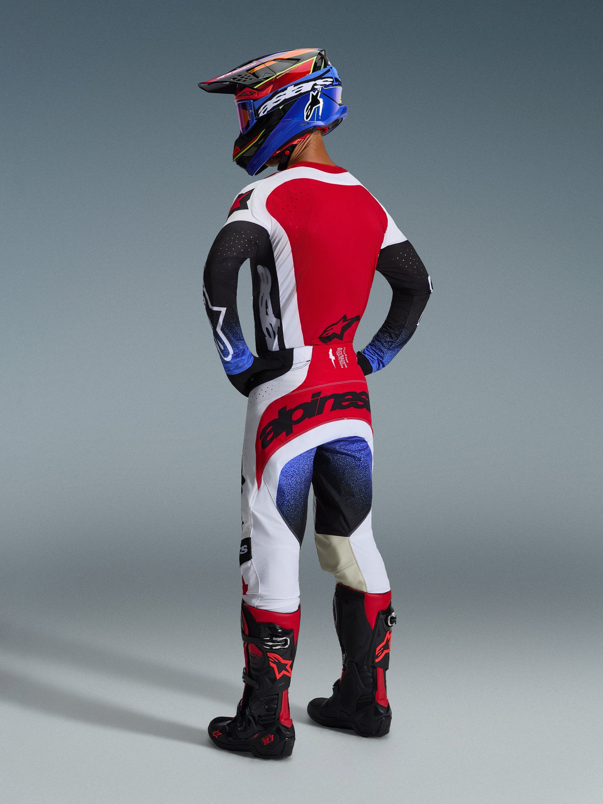 Motocrosskører set bagfra iført komplet off-road udstyr, 2026 Techstar Knif trøje i rød, hvid og sort med blå detaljer på ærmerne, matchende tekniske bukser, blå racerhjelm og sorte og røde støvler, professionelt motocross-racersæt.