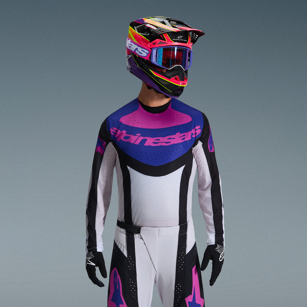 Motocrosskører iført Alpinestars 2026 Techstar Knif udstyrssæt, trøje med blå og lyserød grafik på brystet og hvid torso, hvide bukser med sorte paneler og lyserøde logoer, flerfarvet hjelm i lilla og lyserød med matchende goggles, sorte handsker, professionelt terrænkørselsudstyr