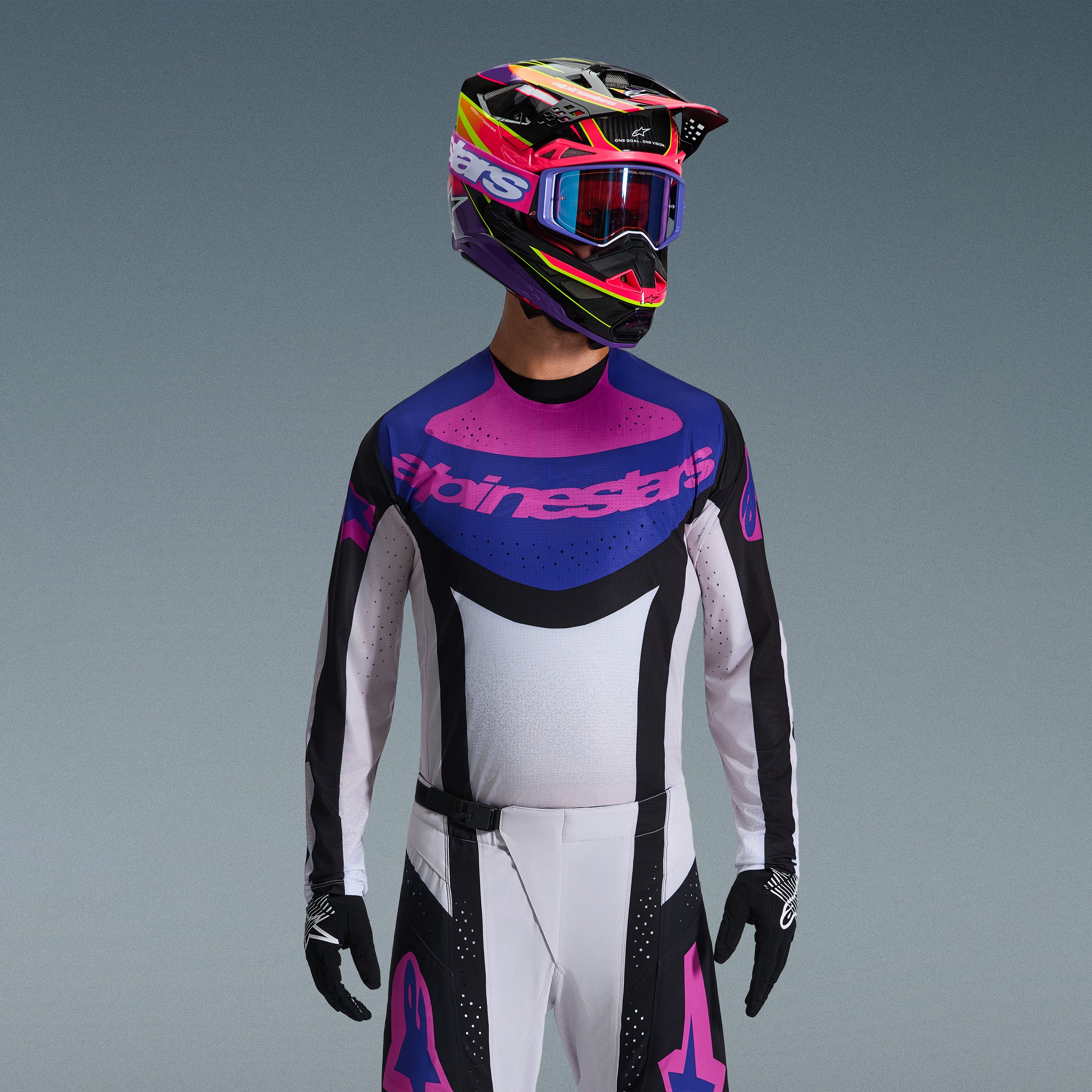Motocrosskører iført Alpinestars 2026 Techstar Knif udstyrssæt, trøje med blå og lyserød grafik på brystet og hvid torso, hvide bukser med sorte paneler og lyserøde logoer, flerfarvet hjelm i lilla og lyserød med matchende goggles, sorte handsker, professionelt terrænkørselsudstyr