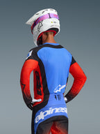 Alpinestars 2026 Techstar Knif Gear Set, motocross-udstyr, kører set bagfra, blå jersey med røde ærmer og sorte detaljer, sorte og blå bukser med hvidt logo, hvid hjelm med lyserød brillerem, laserskårne perforationer til ventilation