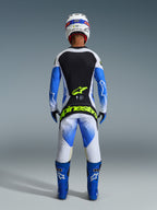 Alpinestars 2026 Techstar Knif-udstyrssæt, set bagfra af en motocross-rytter i Ucla Blue, sort og hvid, med Yellow Fluo-branding på trøjen og bukserne, kombineret med blå racingstøvler og en hvid hjelm.